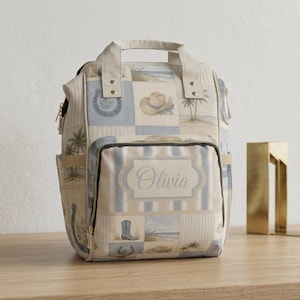 May include: A beige and blue patchwork backpack with a top handle and multiple pockets. The design includes cowboy boots, horseshoes, and beach scenes. The name "Olivia" is displayed on the front pocket.