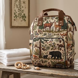 May include: A backpack featuring a cream-colored fabric with a forest-themed print of deer, bears, owls, foxes, and pine trees. The bag includes brown faux leather top handles, a front zippered pocket, and side pockets with a structured rectangular shape.