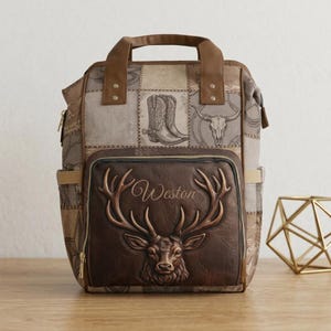 Puede incluir: Una mochila marrón con un bolsillo frontal que presenta una cabeza de ciervo en relieve y el nombre Weston impreso encima. El cuerpo principal tiene un patrón de parches de botas vaqueras, calaveras y estrellas en tonos beige y marrón.