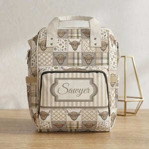 Op de afbeelding: Een beige en bruine rugzak met een patchwork ontwerp met illustraties van Schotse Hooglanders. De voorzak heeft verticale strepen en de naam "Sawyer". De tas heeft een handvat aan de bovenkant en zijzakken.
