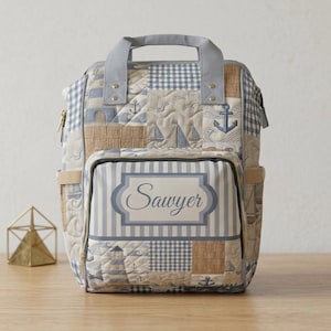 May include: Quilted backpack with a nautical theme. The bag features a patchwork design with anchors, sailboats, and lighthouse motifs in shades of blue, tan, and white. The name "Sawyer" is displayed on the front. It has a gray handle and straps.