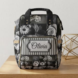 Puede incluir: Mochila con estampado floral en blanco y negro con el nombre "Olivia" en el bolsillo delantero. La mochila tiene un asa superior, correas ajustables y cremalleras doradas. El diseño incluye varios patrones de flores.