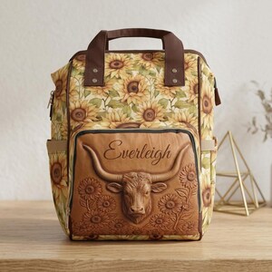 Puede incluir: Una mochila con estampado de girasoles en amarillo, marrón y verde. El bolsillo frontal es de material marrón con una cabeza de buey longhorn y girasoles en relieve. El nombre Everleigh está impreso en cursiva sobre la cabeza del buey.