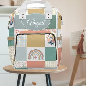 May include: A patchwork-style backpack with a pastel color scheme. The design includes squares of various patterns, including floral prints, a rainbow, and a name in script. The backpack has a top handle and side pockets.