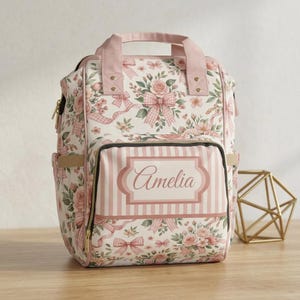 May include: A backpack featuring a white background with a pattern of pink roses, green leaves, and pink gingham bows. The front pocket displays a rectangular label with the name Amelia in cursive script over a vertical pink and white striped background.