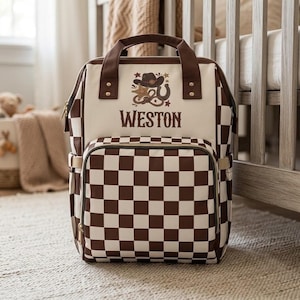 May include: A brown and cream checkered backpack featuring a top handle and a front pocket. The center panel displays the name WESTON in brown lettering above a graphic illustration of a cowboy hat, a sheriff star, a lasso, and a horseshoe.