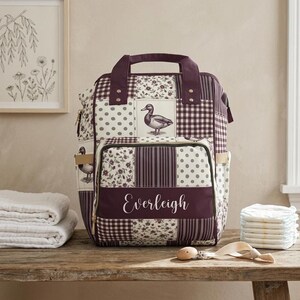 May include: A patchwork backpack featuring a dark plum and cream colour scheme with a repeating pattern of ducks, floral motifs, polka dots, and gingham checks. The front pocket displays the name Everleigh in white script lettering on a plum panel.