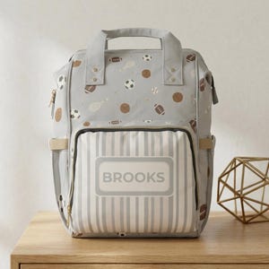 May include: A light gray backpack featuring a repeating pattern of brown footballs, white soccer balls, and tennis rackets. The front pocket has a white and gray vertical striped design with a rectangular label displaying the name BROOKS.