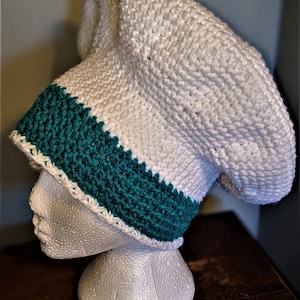 May include: A handmade white crochet hat with a teal band. The hat is displayed on a white foam head. The hat has a textured stitch pattern and a wide brim. The teal band is a solid colour and is located near the bottom of the hat.