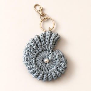 May include: A light blue crocheted nautilus shell keychain with a pearl bead centre. It features a gold-coloured clasp and key ring. The shell is made of yarn.