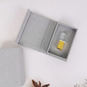 May include: A gray fabric box, open to reveal a gold and clear object. The box is rectangular with a hinged lid. The object has a gold base and a clear, rectangular top. The box and object are on a white surface.