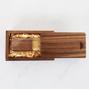 May include: A wooden box with a hinged lid, containing a small wooden USB drive. The box is made of walnut wood and has a natural finish. The USB drive is nestled in shredded paper.
