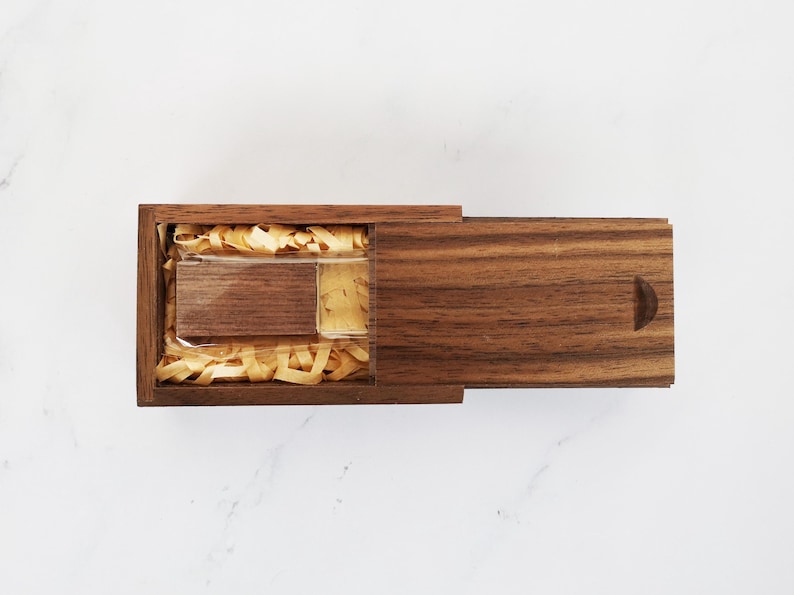 May include: A wooden box with a hinged lid, containing a small wooden USB drive. The box is made of walnut wood and has a natural finish. The USB drive is nestled in shredded paper.