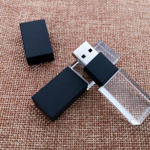 Puede incluir: Una memoria USB negra con una carcasa de cristal transparente. La unidad está en posición cerrada, con el conector USB oculto dentro de la carcasa.