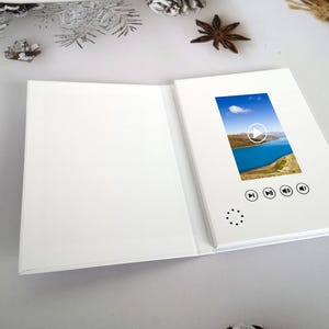 May include: A white video brochure, open to display a screen with a landscape image of a lake and mountains. The screen has play, rewind, and volume control icons. The brochure is on a white surface.