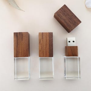 Puede incluir: Tres unidades flash USB con una base rectangular transparente y una parte superior de madera. Una unidad tiene el conector USB expuesto. Las partes superiores de madera son de color marrón cálido. Las unidades están dispuestas sobre una superficie clara.