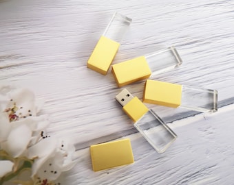 May include: Five gold and clear crystal USB flash drives on a white wooden surface. The flash drives have a gold metal top and a clear crystal body.