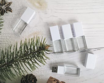 Könnte beinhalten: Fünf weiße USB-Sticks mit durchsichtigen Kunststoffgehäusen auf einer weißen Holzoberfläche. Ein kleiner Kiefernzweig und eine Kiefernzapfen befinden sich in der linken unteren Ecke des Bildes.