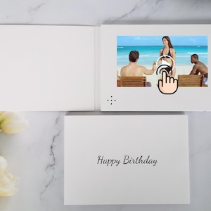 Può includere: Un biglietto d'auguri video bianco con uno schermo che mostra una scena di spiaggia con tre persone. Il biglietto è aperto, mostrando una griglia dell'altoparlante, e un biglietto chiuso sottostante mostra il testo nero Happy Birthday in corsivo.