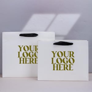 Può includere: Due borse della spesa bianche con manici neri. Le borse hanno la scritta "YOUR LOGO HERE" stampata in colore oro. Le borse sono su una superficie bianca con sfondo grigio.