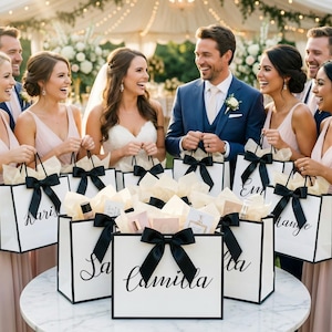 May include: A group of people, including a bride and groom, are smiling and holding white gift bags with black bows. The bags have black trim and names written on them. The bags are filled with gifts and tissue paper.