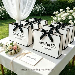 May include: White gift bags with black trim and black bows, personalized with the name "Sandra" and the word "Bridesmaid". A bouquet of flowers and a welcome card are on the table. The bags are arranged on a white table.