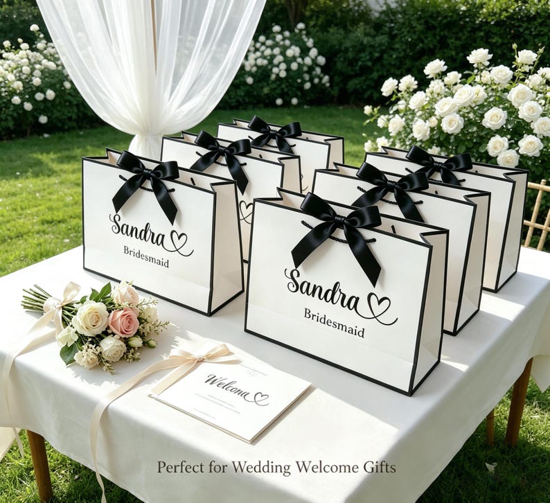 May include: White gift bags with black trim and black bows, personalized with the name "Sandra" and the word "Bridesmaid". A bouquet of flowers and a welcome card are on the table. The bags are arranged on a white table.