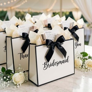 May include: White gift bags with black trim and bows, filled with tissue paper and beauty products. The bags are labeled with the words "Thank You", "Bridesmaid", and "You". A white rose and baby's breath are in the foreground.