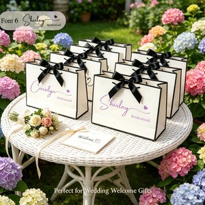 May include: A collection of white gift bags with black trim and black bows, arranged on a white wicker table. Each bag is personalized with a name in purple script and the word "Bridesmaid". A small bouquet and welcome card are also present.