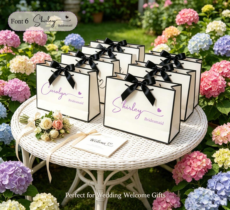 May include: A collection of white gift bags with black trim and black bows, arranged on a white wicker table. Each bag is personalized with a name in purple script and the word "Bridesmaid". A small bouquet and welcome card are also present.
