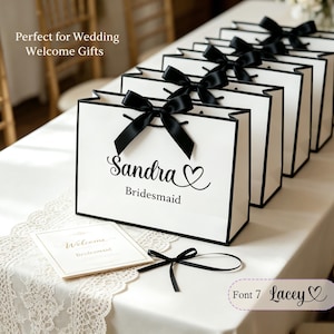 May include: White gift bags with black trim and black satin bows. The bags are personalized with the name "Sandra" and the word "Bridesmaid" in black script. A small card and a black bow are also on the table.