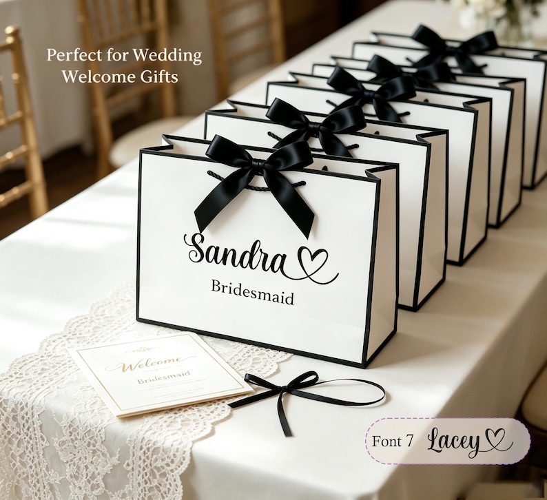 May include: White gift bags with black trim and black satin bows. The bags are personalized with the name "Sandra" and the word "Bridesmaid" in black script. A small card and a black bow are also on the table.