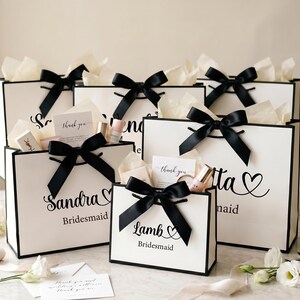 May include: White gift bags with black trim and black bows, filled with gifts and tissue paper. The bags are labeled with names and the word "Bridesmaid". Small cards and beauty products are visible.