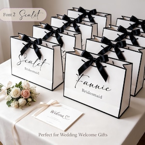 May include: White gift bags with black trim and black satin bows. The bags are personalized with names in elegant script, with the word "Bridesmaid" below. A small bouquet of flowers and a welcome card are also present.
