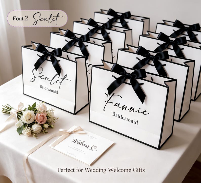 May include: White gift bags with black trim and black satin bows. The bags are personalized with names in elegant script, with the word "Bridesmaid" below. A small bouquet of flowers and a welcome card are also present.
