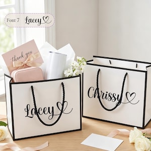 May include: Two white gift bags with black trim and rope handles. One bag says "Lacey" with a heart, the other says "Chrissy" with a heart. The "Lacey" bag contains a pink card, jewelry box, and a white tumbler.