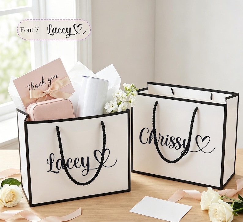 May include: Two white gift bags with black trim and rope handles. One bag says "Lacey" with a heart, the other says "Chrissy" with a heart. The "Lacey" bag contains a pink card, jewelry box, and a white tumbler.