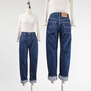 May include: Two mannequins display a pair of dark wash denim jeans with a white long-sleeve shirt. The jeans have a classic fit, with a straight leg and a cuffed hem. The jeans have a leather patch on the back pocket.