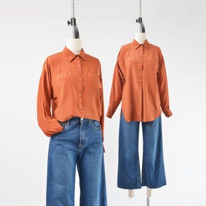May include: Two mannequins display a burnt orange long-sleeve shirt with a button-down front, paired with blue denim wide-leg jeans. The shirts have a subtle sheen, and the jeans have a classic, relaxed fit. The mannequins are set against a plain white background.