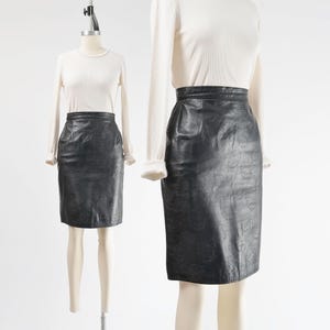 May include: A black leather pencil skirt displayed on a mannequin. The skirt is knee-length and has a high waist. The skirt is paired with a white long-sleeve top. The skirt appears to be made of a textured leather material.