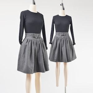 May include: Two mannequins display a black long-sleeve top paired with a gray pleated skirt. The skirt features a belt with a buckle. The outfits are presented against a white background, showcasing the classic design.
