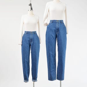 Puede incluir: Dos maniquíes muestran camisetas blancas de manga larga con cuello redondo combinadas con vaqueros azules de tiro alto. Los vaqueros presentan un diseño clásico de cinco bolsillos, cierre de botones y corte recto. Un par tiene el bajo doblado.