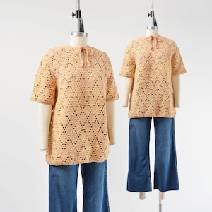 May include: Two peach-coloured, short-sleeved crochet tops with a diamond pattern are displayed on mannequins. The tops have a tie at the neckline and are paired with blue denim wide-leg trousers. The mannequins stand against a white background.
