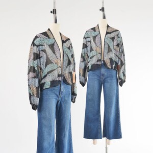 May include: Two open-front jackets with a leaf pattern in pastel colors and black sequins are displayed on mannequins, paired with blue denim wide-leg jeans. The jackets have long sleeves and are styled for a vintage aesthetic.