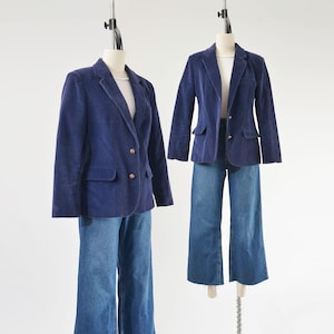 May include: Two navy blue corduroy blazers with gold buttons and matching blue denim jeans are displayed on mannequins. The blazers have a classic design with notched lapels and front pockets. The jeans are wide-legged.