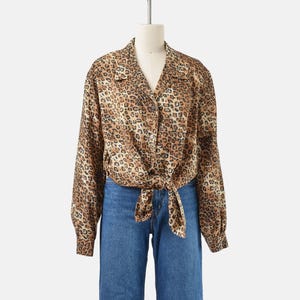 May include: A button-down leopard print blouse with long sleeves and a knotted front. The blouse is displayed on a mannequin with blue jeans. The blouse features a classic collar and a V-neckline. The leopard print is brown and black.