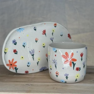 Può includere: Una tazza in ceramica e un piatto ovale abbinato, entrambi caratterizzati da uno smalto bianco sporco maculato decorato con piccoli fiori dipinti a mano nei toni dell'arancione, giallo, blu, viola e rosso, con steli e foglie verdi.