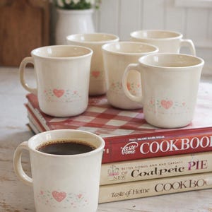 May include: Cream-colored ceramic mugs with heart and floral designs. One mug is filled with coffee. The mugs are stacked on cookbooks with red and white checkered fabric. A vase of white flowers is in the background.