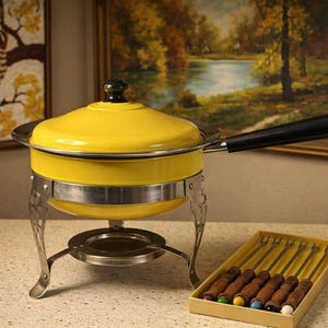 May include: A vintage yellow fondue pot with a black handle and lid. The pot sits on a silver stand with decorative legs. A box of fondue forks with wooden handles and colored tips is next to the pot.