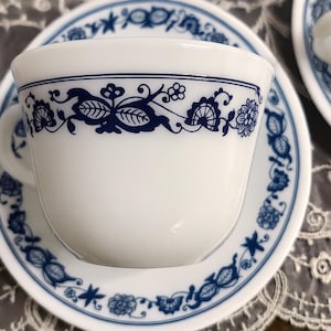 May include: White teacup and saucer set with a blue floral design. The cup and saucer have a repeating pattern of flowers, leaves, and other decorative elements. The teacup has a handle and sits on a matching saucer.